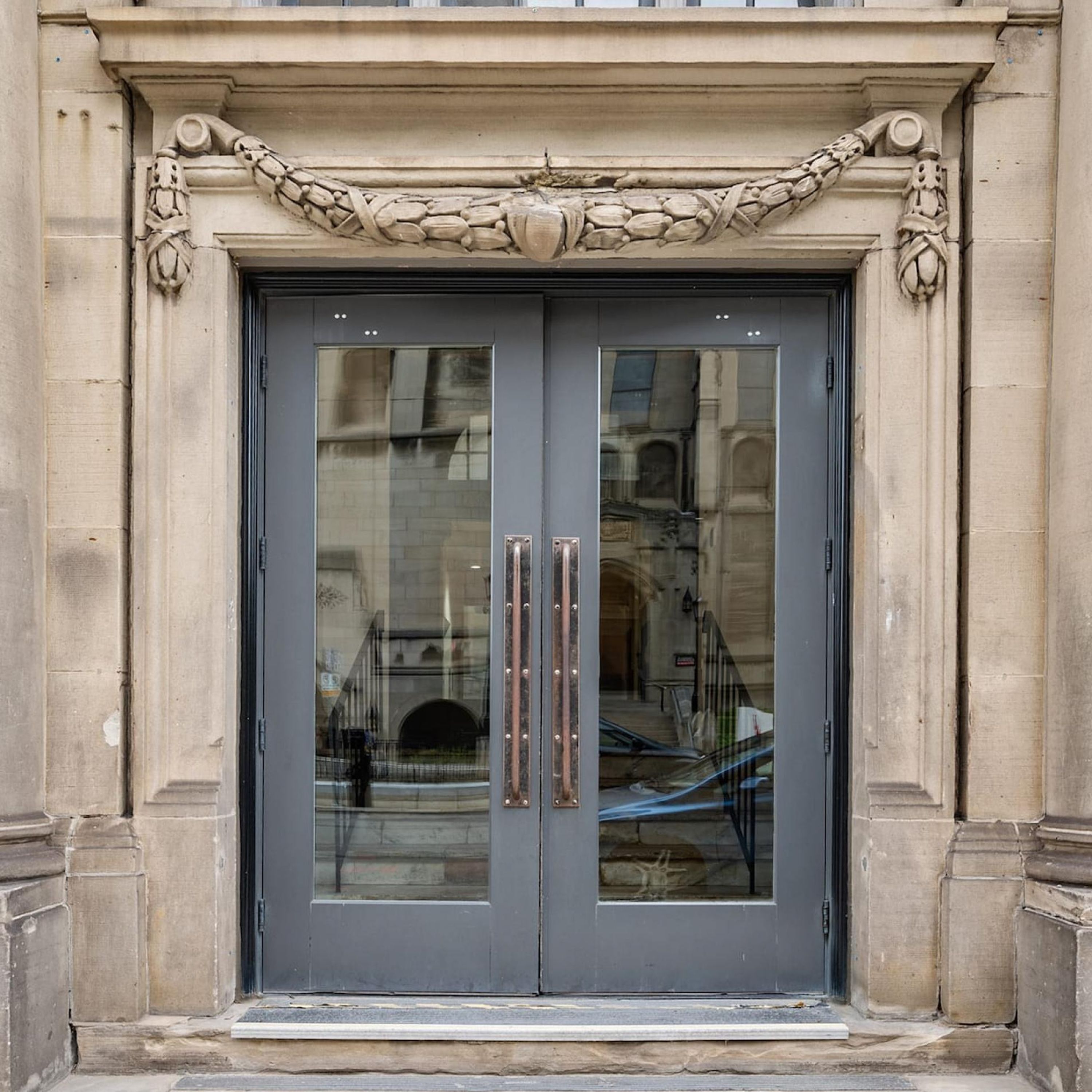 Une porte à deux battants ornée d’un panneau de verre réfléchissant, flanquée de colonnes en pierre et d’une frise décorative.