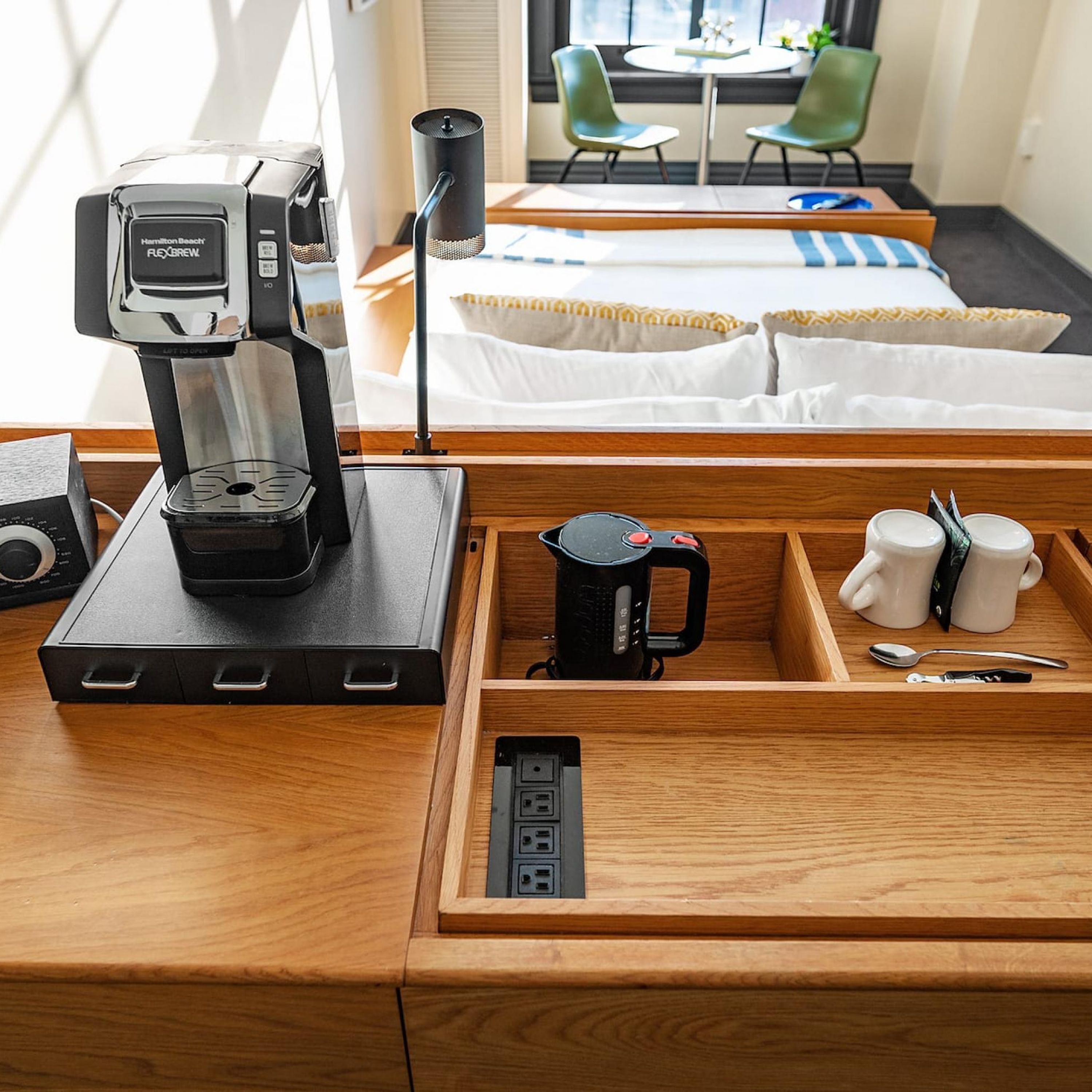 A hotel room with a wooden desk, a coffee maker, a mug, a spoon, and a power outlet.