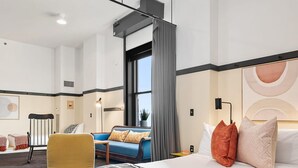 A modern hotel room with a large bed, a sofa, a yellow armchair, and a view of the city through a window. - Kasa Family Space Historic Ymca The Maverick (Pittsburgh)