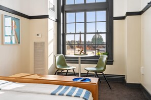 A hotel room with a bed, a small table with chairs, a window with a view of a building, and a painting on the wall. - Kasa See a Show at Kelly Strayhorn Theater The Maverick (Pittsburgh)