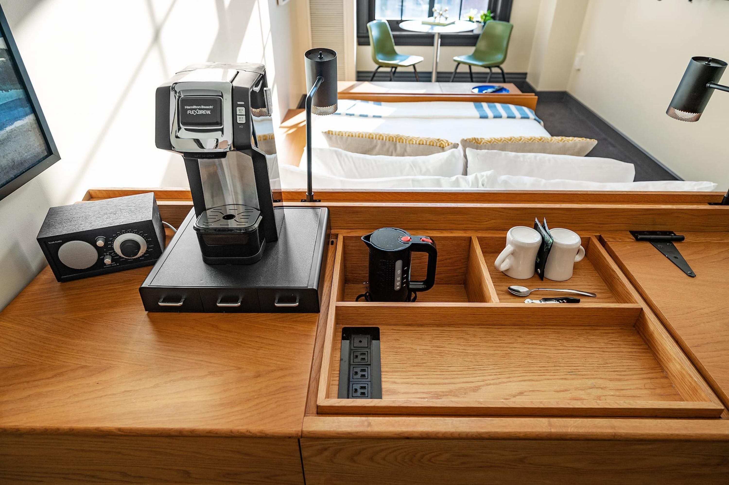 Ein Hotelzimmer mit Holzschreibtisch, einer Kaffeemaschine, einem Radio, einer Ablage mit Tassen und Besteck sowie einem Bett mit weißer Bettwäsche.