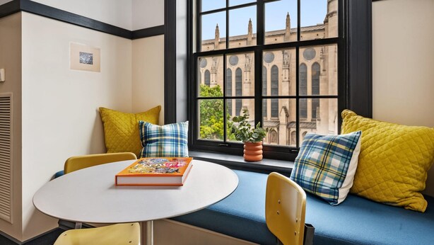 A cozy seating area with a round table, yellow chairs, and a blue bench with plaid cushions. A book rests on the table, and a potted plant is on the bench. - Kasa Enjoy Local Dining in East Liberty The Maverick (Pittsburgh)