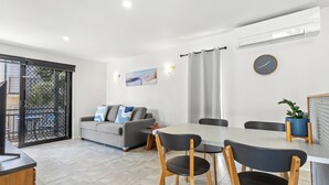 A modern living room with a grey sofa, a dining table with four chairs, a potted plant, and a wall clock. - Wake up each morning just steps away from 7-mile Beach and the iconic boardwalk (Lennox Head)