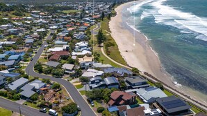 An aerial view of a coastal residential area with houses, a beach, and a road. - Wake up each morning just steps away from 7-mile Beach and the iconic boardwalk (Lennox Head)