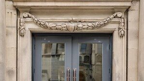 Une porte à deux battants ornée d’un panneau de verre réfléchissant, flanquée de colonnes en pierre et d’une frise décorative.