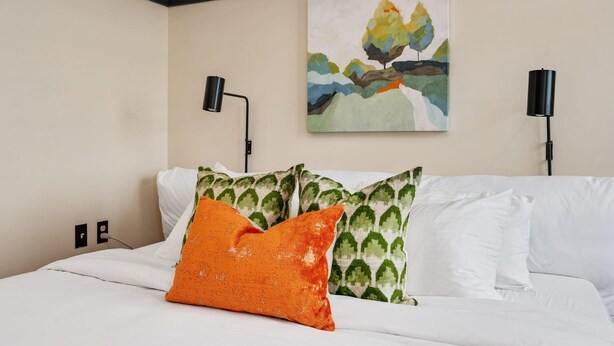 Une chambre d’hôtel avec un lit, du linge de lit blanc, des coussins à motifs verts, un coussin orange, un tableau représentant un arbre accroché au mur et deux lampes murales noires.