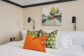 A hotel room with a bed, white bedding, green patterned pillows, an orange pillow, a painting of a tree on the wall, and two black wall lamps. - Kasa Walk to Bakery Square Boutique Studio The Maverick (Pittsburgh)