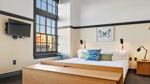 Une chambre moderne avec un grand lit, un téléviseur fixé au mur, une fenêtre donnant sur des immeubles et une œuvre d’art représentant des papillons accrochée au mur.