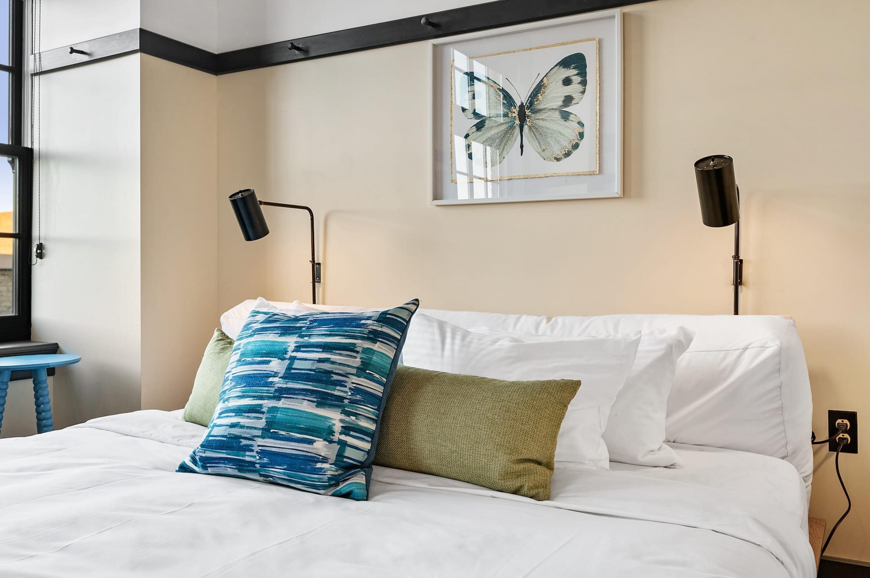 A neatly made bed with white linens, two pillows, a framed butterfly artwork on the wall, and two black desk lamps.