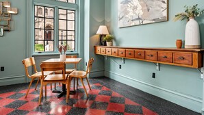 Une pièce avec un sol à carreaux, une table en bois entourée de chaises, une fenêtre donnant sur des immeubles, une étagère fixée au mur avec une lampe, et un tableau.
