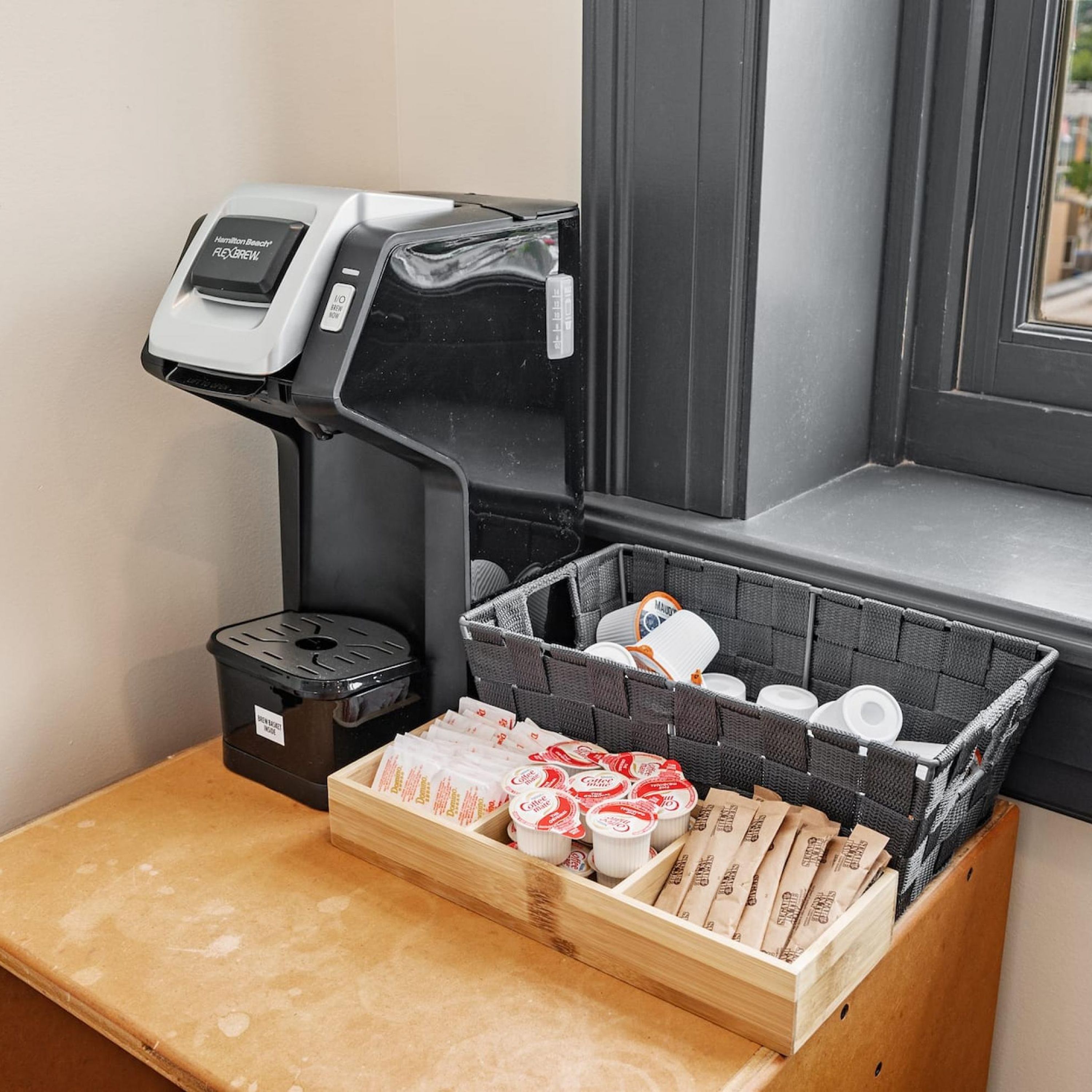 Une machine à café, une poubelle et un panier contenant des capsules de café, ainsi qu’un plateau en bois, sont posés sur un comptoir.