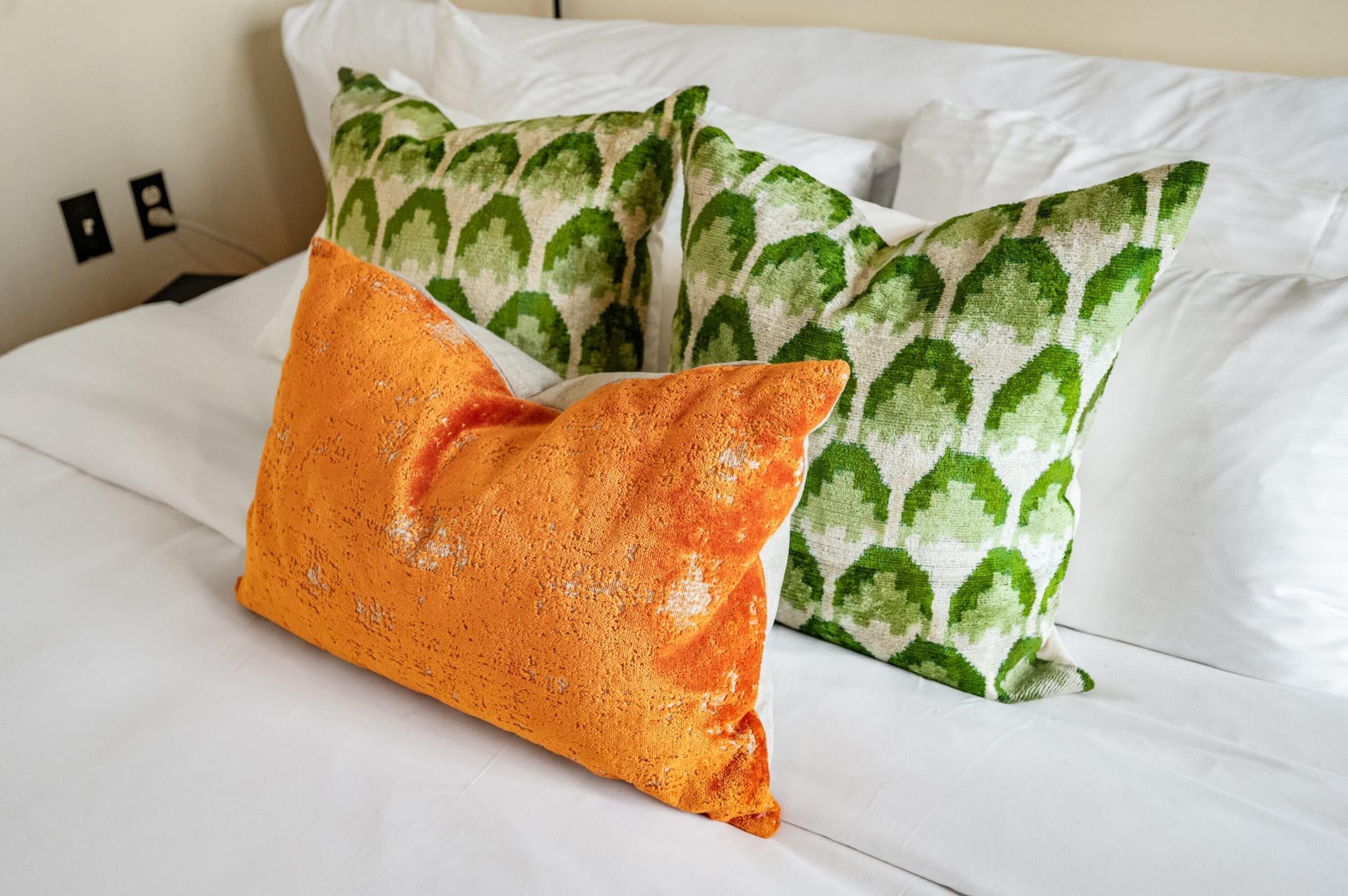 A bed with white bedding and two pillows, one orange and one green with a leaf pattern.