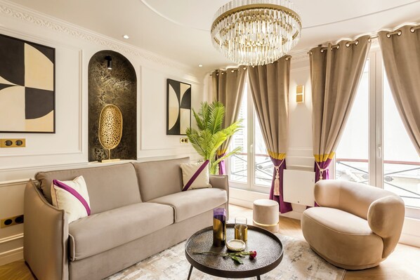 A modern living room with a beige sofa, a round coffee table, and a decorative chair. There is a large window with curtains, a chandelier, and wall art.