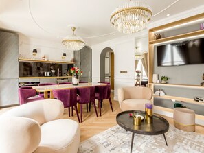 Ein modernes Wohnzimmer mit einem мраморный Couchtisch, bequemen Sitzmöglichkeiten, einem Kronleuchter und einem an der Wand montierten Flachbildfernseher.