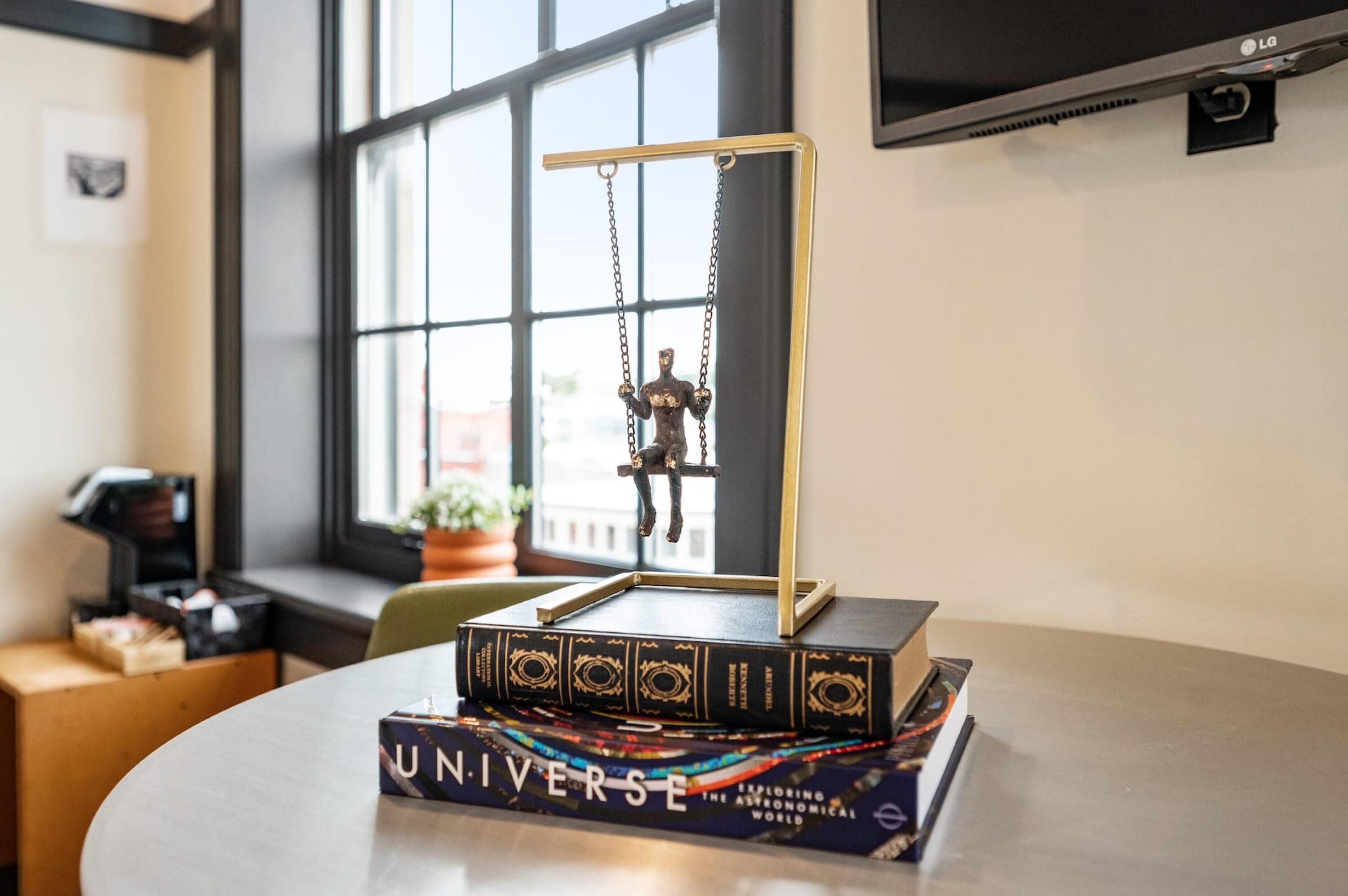 A table with a decorative metal sculpture, a stack of books, and a television mounted on the wall.