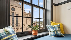 A room with a large window overlooking a historic building, a blue bed with plaid pillows, and a potted plant on the windowsill.