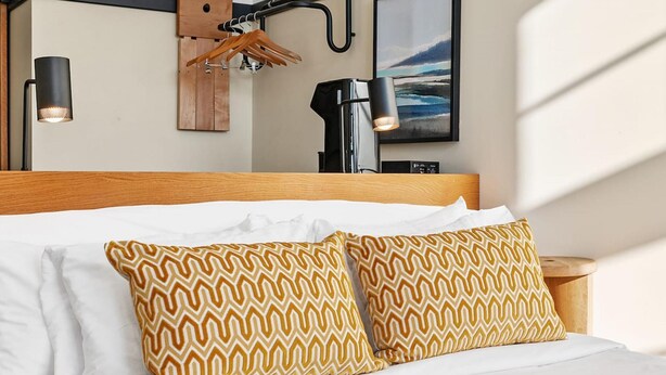 A neatly made bed with white linens and patterned pillows, a wooden headboard, a wall-mounted lamp, and a framed picture of a landscape. - Kasa Walk to Kelly Strayhorn Theater The Maverick (Pittsburgh)