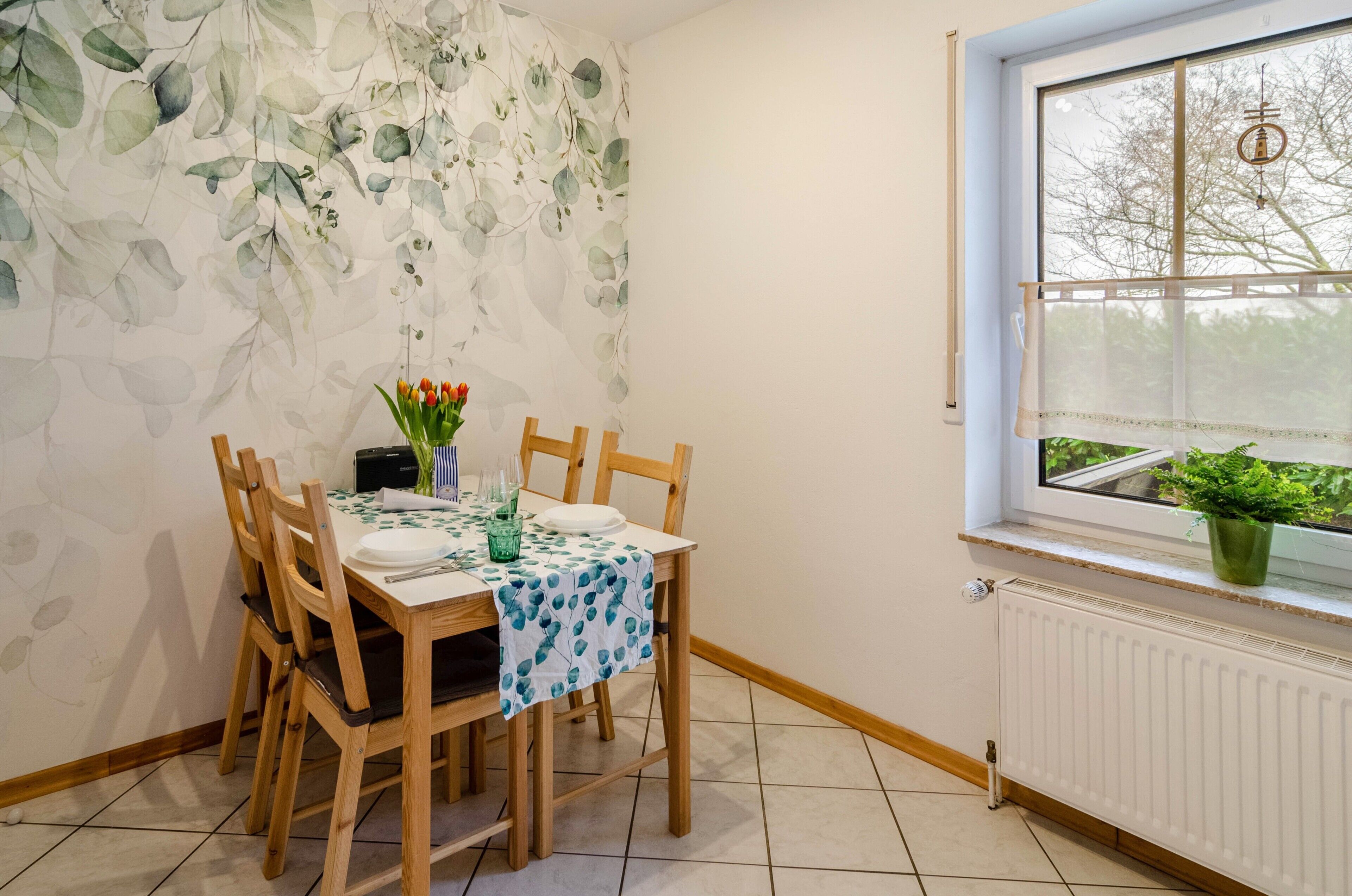 A dining area with a table set for two, a window with a view of greenery, and a radiator below.
