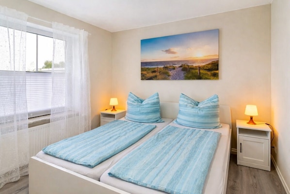 A double bed with matching pillows, two bedside tables with lamps, a window with sheer curtains, and a landscape painting on the wall.