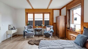 A room with a bed, two armchairs, a small table, a wooden wardrobe, and a window with blinds.