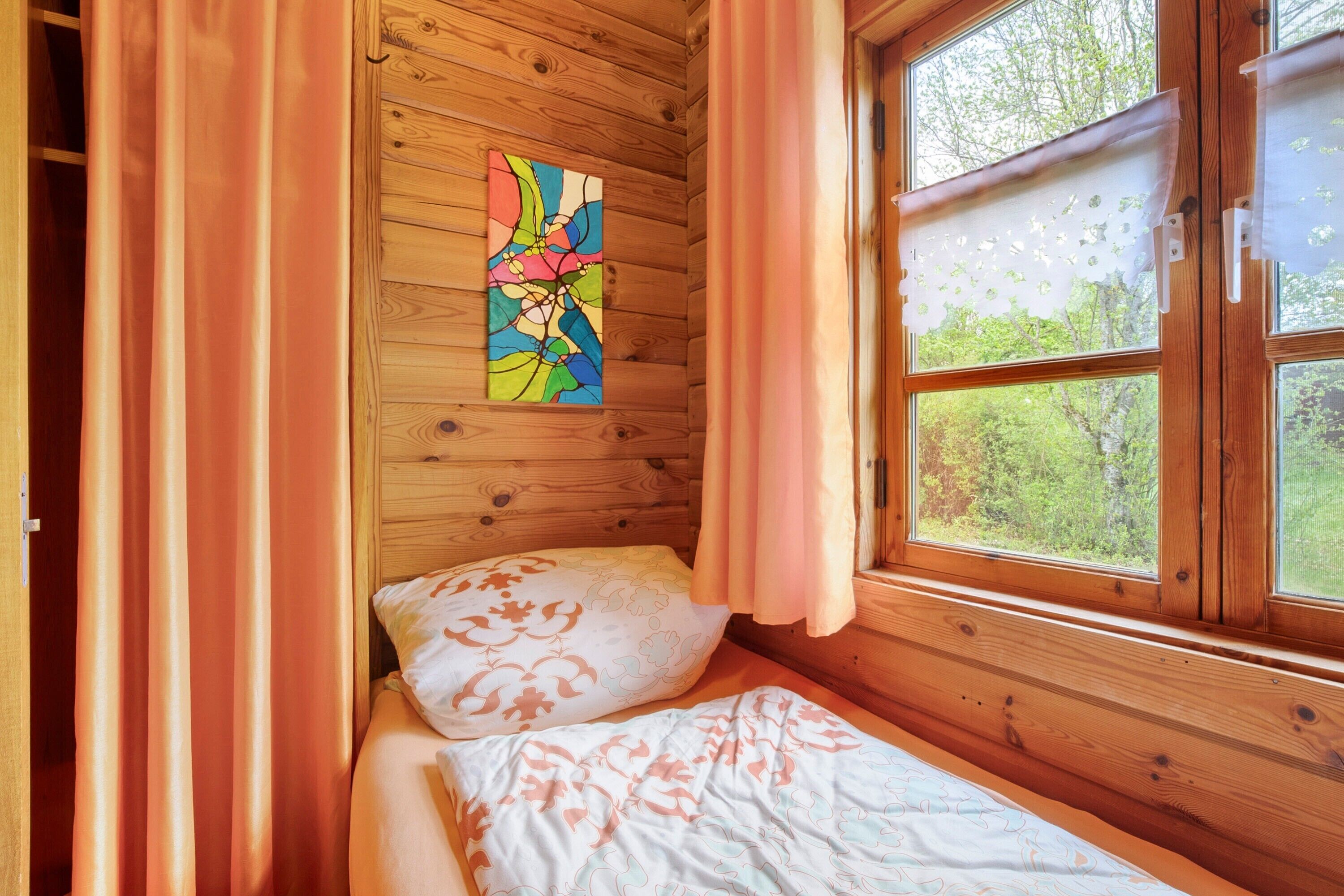 A wooden cabin room with a bed, curtains, and a window with a view of trees.