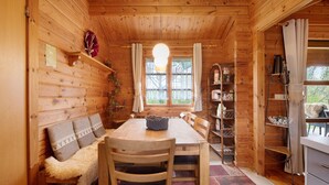 A wooden cabin interior with a dining table, chairs, a bench with cushions, a hanging light fixture, a window with curtains, and shelves with various items. - Holiday Home 'Hüssli Numero 1' with Private Terrace, Shared Garden and Wi-Fi (Hayingen)
