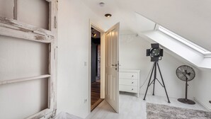 A room with a sloped ceiling, a white door, a small white dresser, a tripod-mounted projector, and a standing fan.