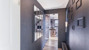 A hallway with a dark wall, a wooden floor, and a staircase leading to a room with a brick wall and a fireplace.