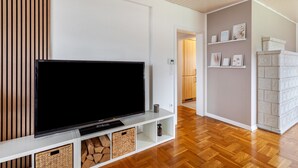 Ein modernes Wohnzimmer mit einem Flachbildfernseher auf einer weißen Unterhaltungsanlage, Holzböden und einer im Hintergrund sichtbaren Küche.
