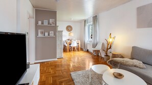 Ein modernes Wohnzimmer mit Flachbildfernseher, einem grauen Sofa, einem weißen Couchtisch und einem Essbereich mit Holztisch und Stühlen.