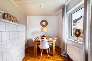 Ruokailutila, jossa on puinen pöytä, valkoiset tuolit, riippuvalaisin ja ikkuna, jossa on verhot.