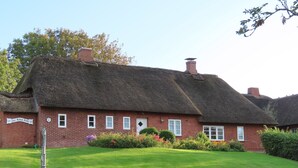 Ett traditionellt hus med halmtak och en fasad av tegel, med husnummer ’2’, och en välskött gräsmatta.