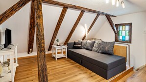 Dormitorio tipo loft con vigas de madera a la vista, un sofá gris, una mesita de noche blanca pequeña y un televisor sobre un soporte blanco.