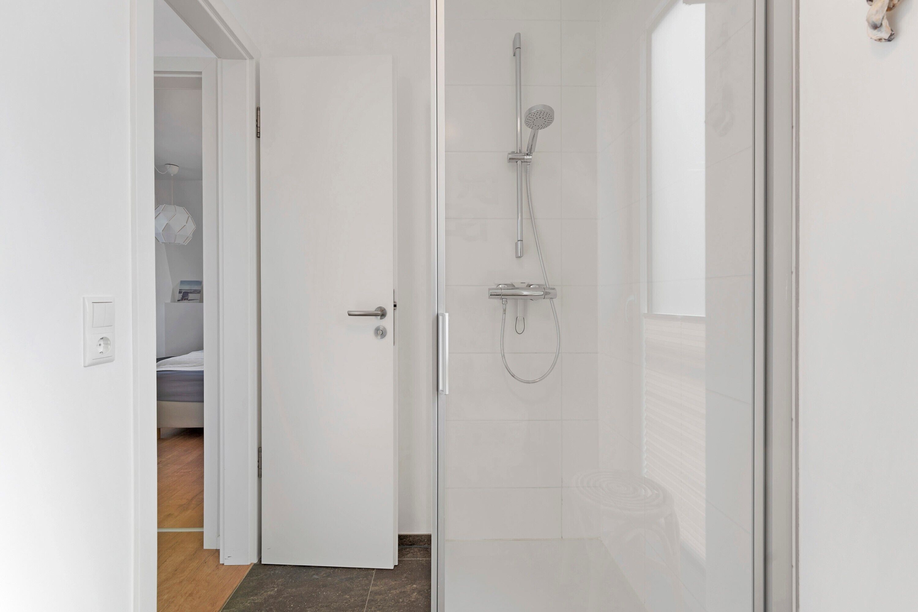 A modern bathroom with a glass shower enclosure, a visible showerhead, and a door leading to a bedroom.