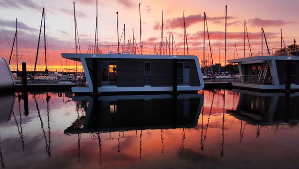 A marina with several boats docked, a modern building with large windows, and a sunset in the background. - Houseboat 'Sutje' with Sea View, Private Terrace and Wi-Fi (Großenbrode)