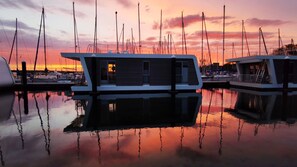 A marina with several boats docked, a modern building with large windows, and a sunset in the background.