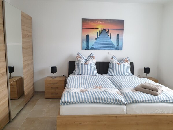 Une chambre à coucher comprenant un lit, deux tables de chevet, une commode, un miroir et un tableau représentant un coucher de soleil sur une jetée.