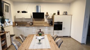 A modern kitchen with white cabinets, a wooden dining table, and a TV mounted above the counter.