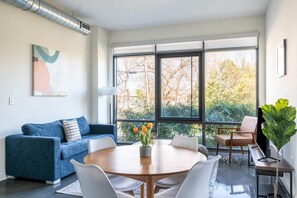 A modern living room with a blue sofa, white chairs, a wooden dining table, and a large window with a view of trees. - Kasa Charming 2 BD Fire Pit Alexandria (Alexandria)