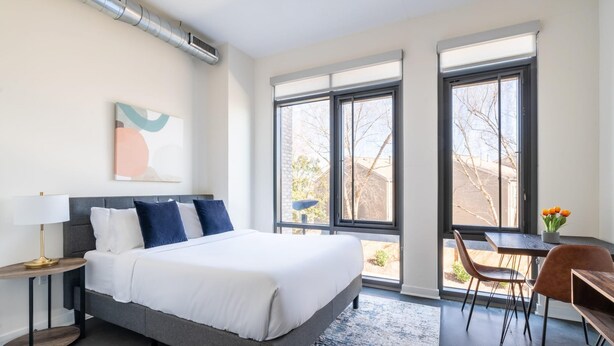 A modern bedroom with a large bed, a desk, and a chair. There are large windows and a view of the outside. - Kasa Cozy Studio Amenity Access Alexandria (Alexandria)