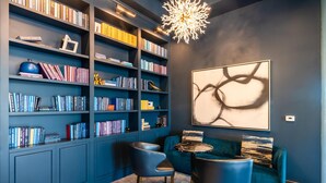 A modern library with dark blue walls, a large bookshelf filled with books, a comfortable blue sofa, and a unique abstract painting. - Kasa Cozy Studio Amenity Access Alexandria (Alexandria)
