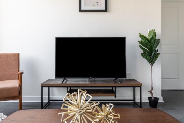 Una sala de estar con un televisor de pantalla plana sobre un soporte de madera, una planta en maceta y una mesa de centro con objetos decorativos.