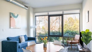 A modern living room with a blue sofa, white chairs, a wooden dining table, and a large window with a view of trees. - Kasa Comfy 2 BD Gym Access Alexandria (Alexandria)