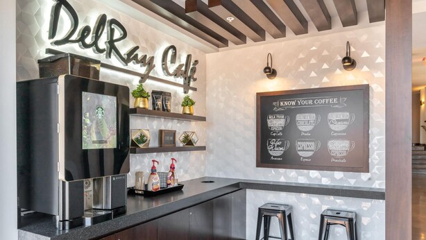 A modern coffee bar with a sign reading "DelRay Cafe," a coffee machine, bar stools, and a menu board displaying coffee options. - Kasa Comfy 2 BD Gym Access Alexandria (Alexandria)