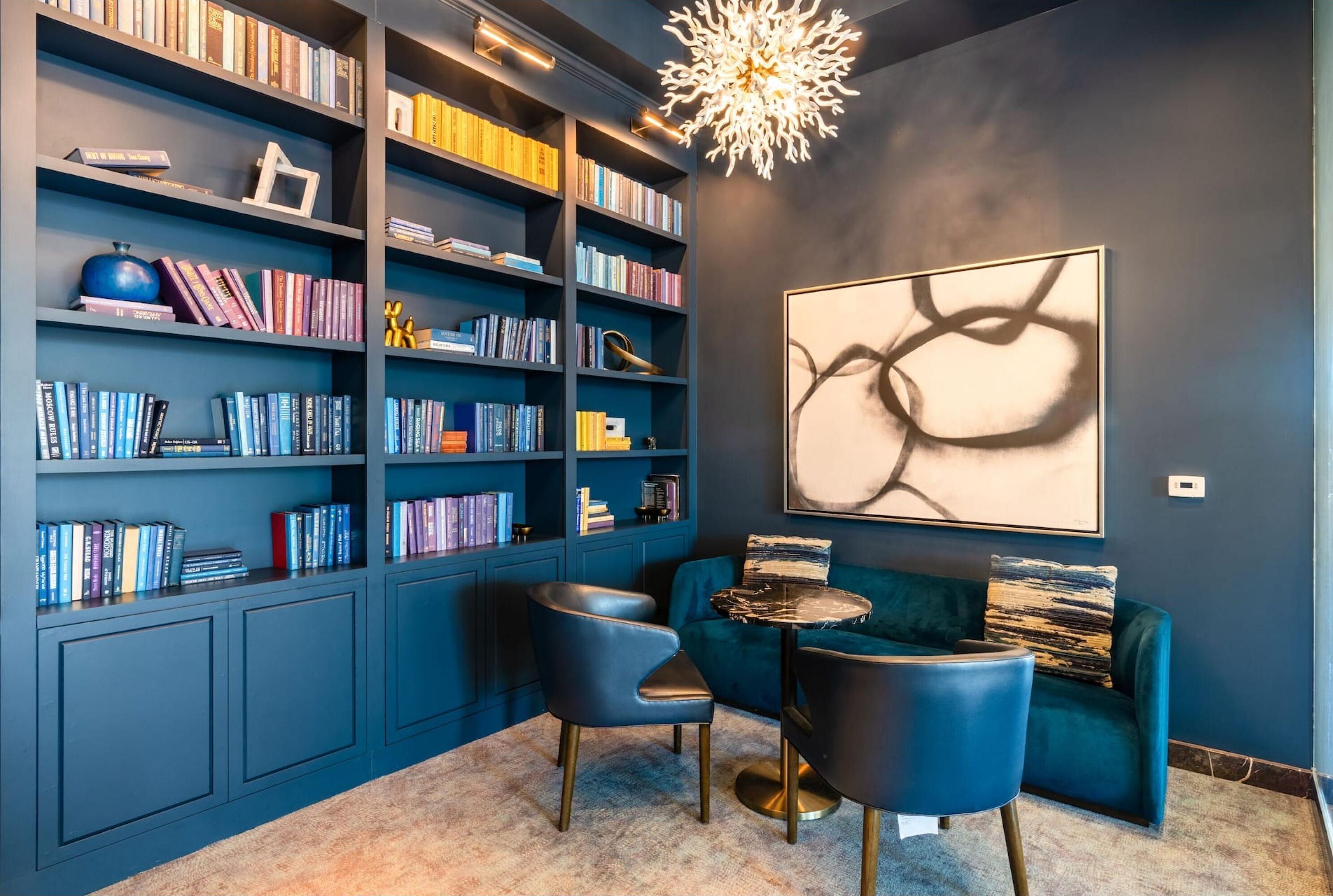 Una biblioteca moderna con paredes azul oscuro, una estantería grande, un sofá azul y un cuadro abstracto único.