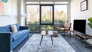 A modern living room with a blue sofa, a coffee table, a TV, and large windows offering a view of trees. - Kasa Comfy 2 BD Gym Access Alexandria (Alexandria)