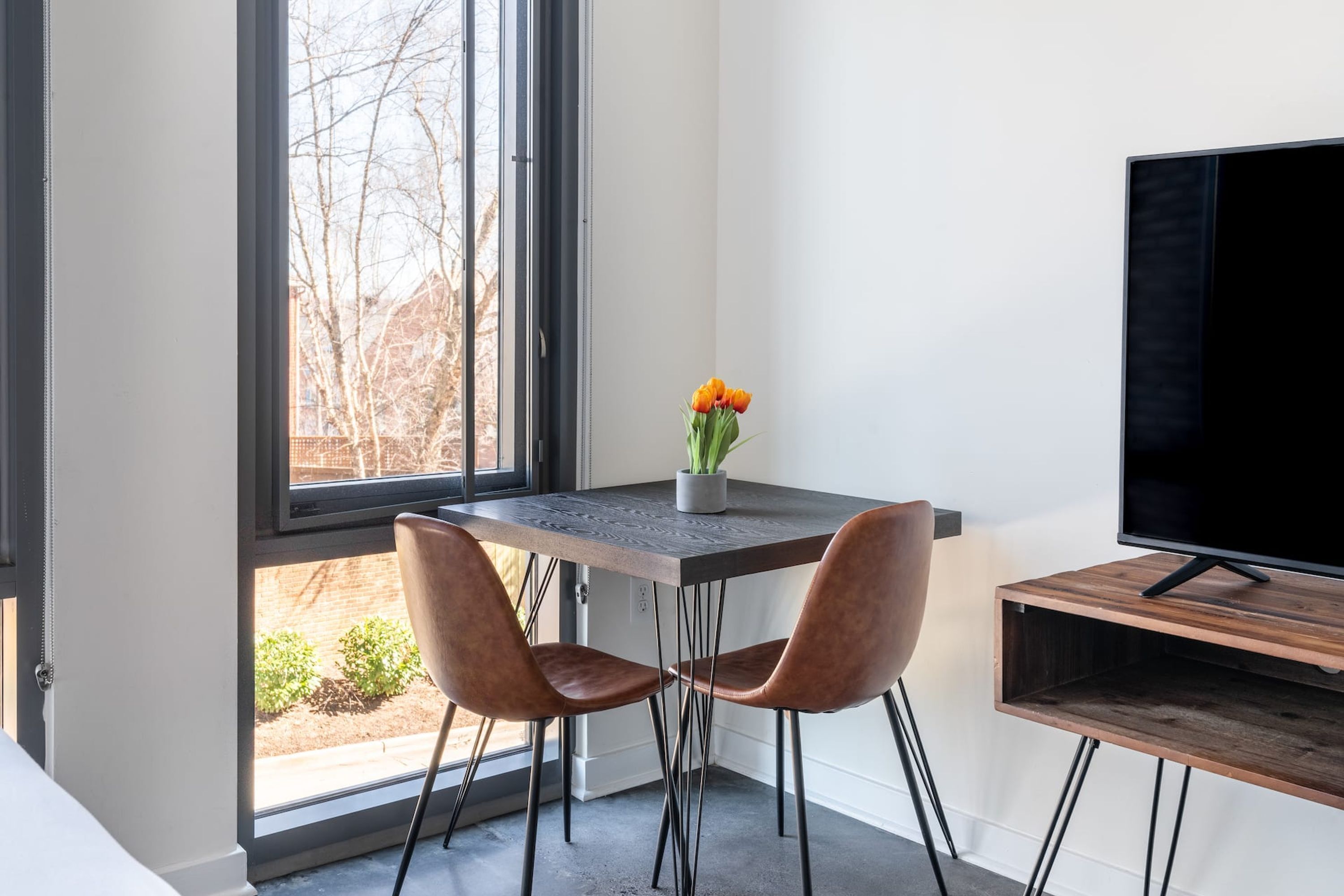 Een moderne kamer met een groot raam, een houten tafel met twee stoelen, een kleine potplant en een flatscreen televisie op een houten voet.