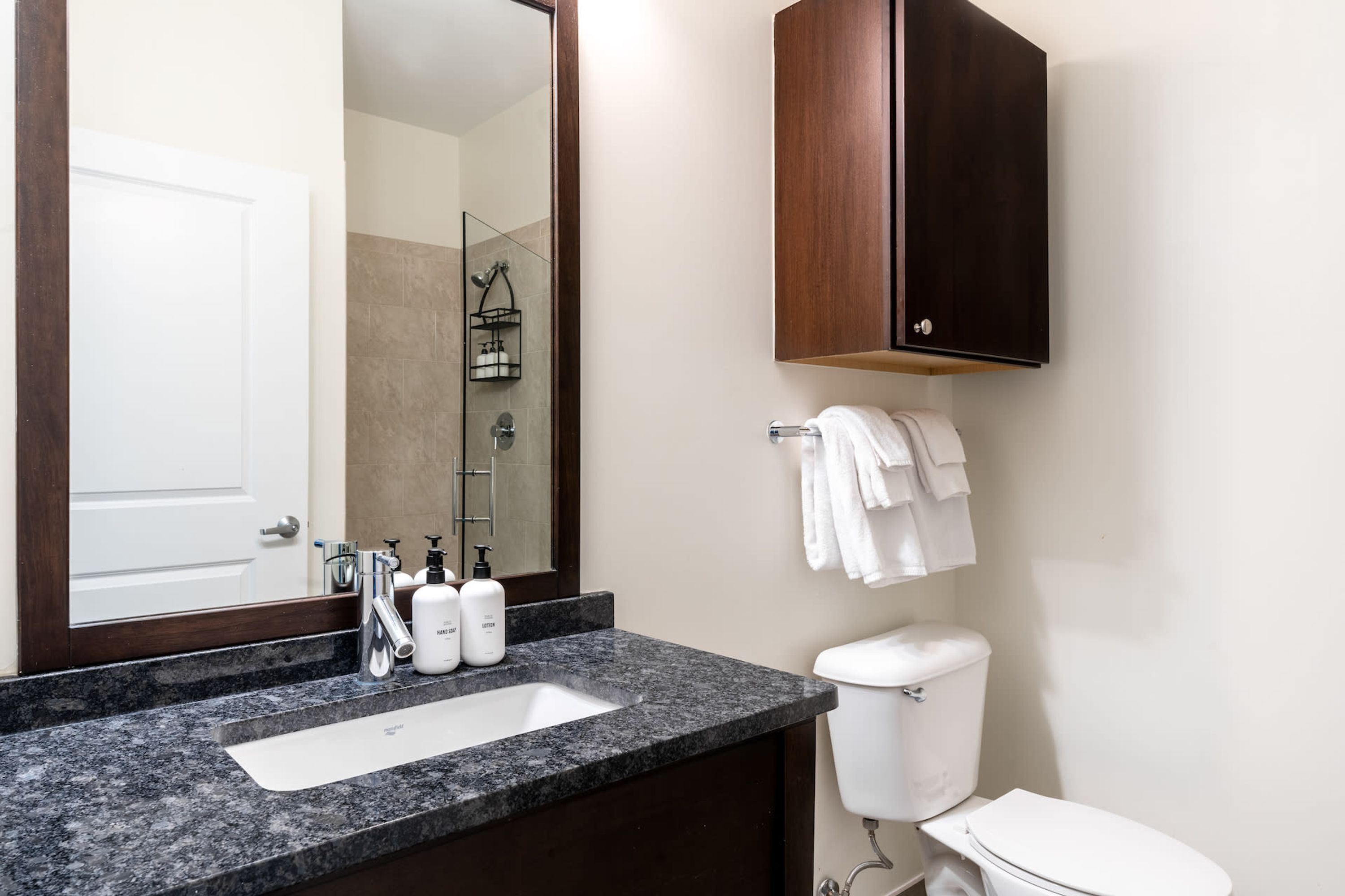 A bathroom with a granite countertop sink, a mirror, a toilet, and a wooden cabinet above the toilet.