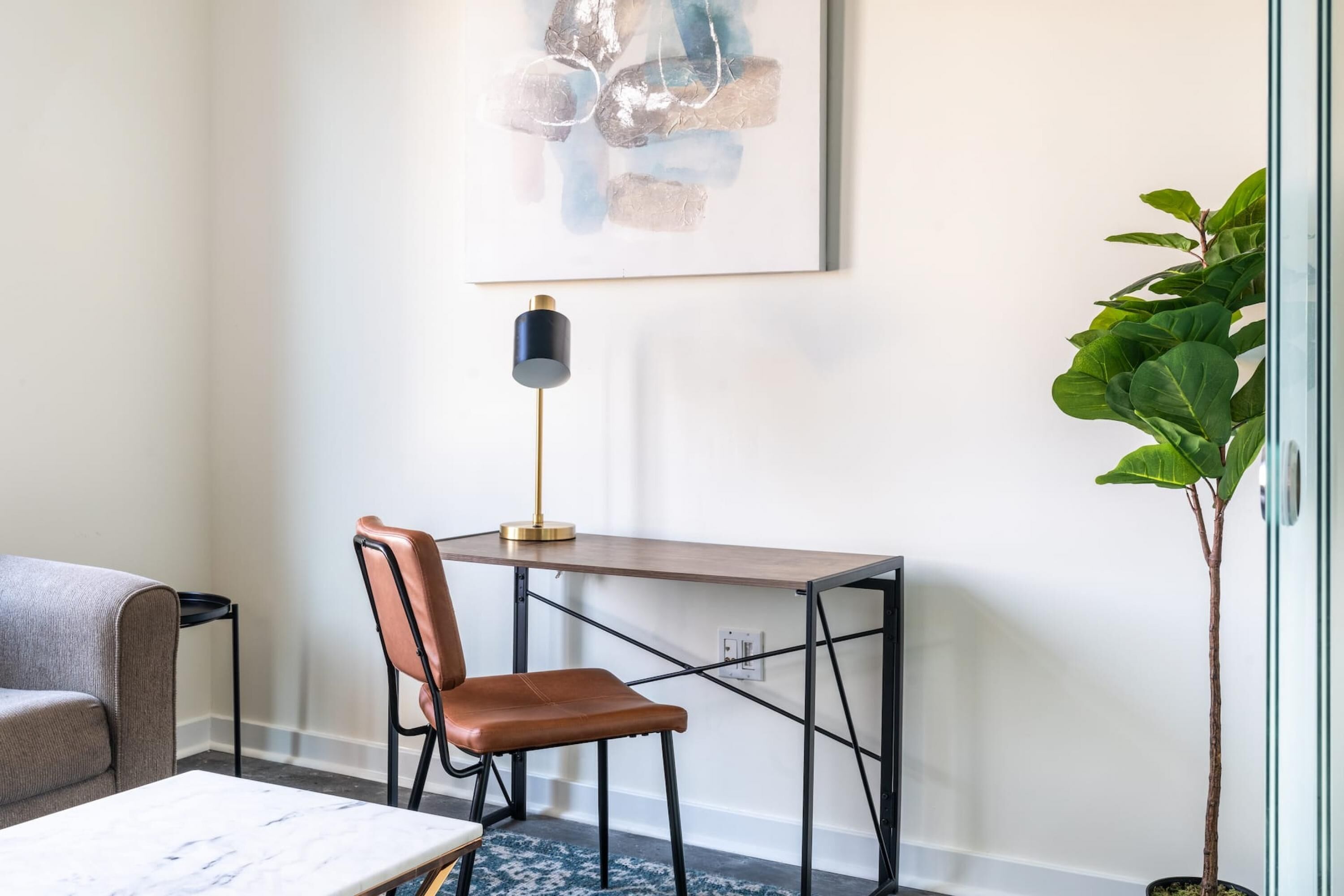 A modern room with a marble table, a brown leather chair, a desk with a lamp, and a large green plant.