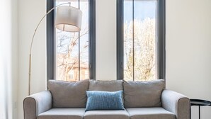 Een moderne woonkamer met een grijze bank, een blauw kussen, een vloerlamp en uitzicht op bomen door grote ramen.